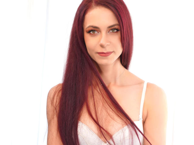 CindeFunRella has long red hair, green eyes, and is wearing a white bra. Her tits are small and her ass is not visible. She has a slight smile.
