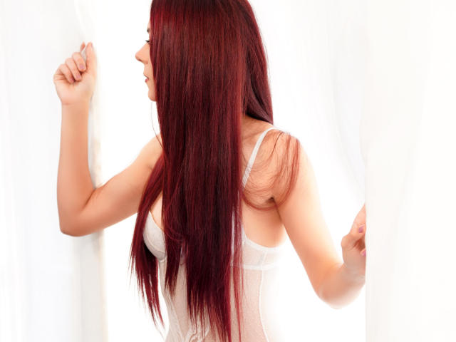 CindeFunRella with long red hair, white lace lingerie, and a side profile, holding white curtains, showing her tits and a cute smile.