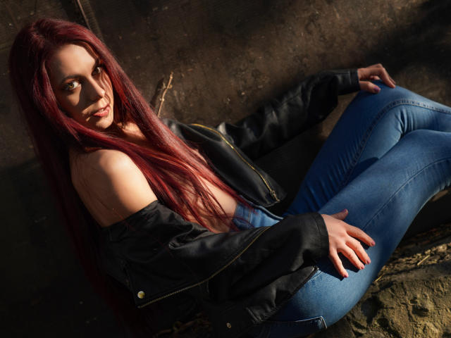 CindeFunRella with red hair, black jacket, and blue jeans, sitting on a rock, looking back with a smile, showing her bare shoulder and hand on her ass.