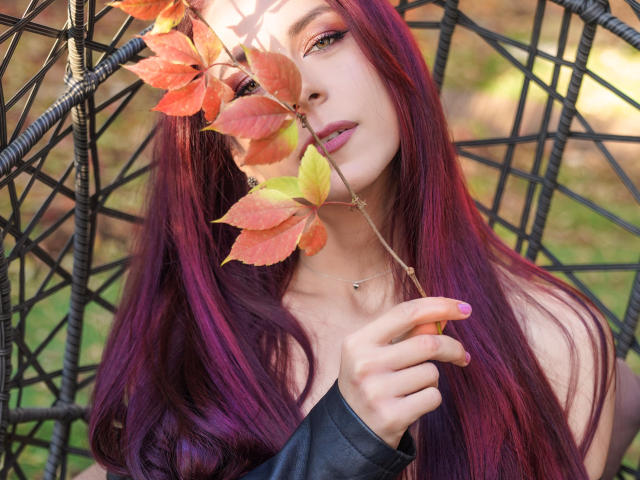 CindeFunRella with purple hair holding autumn leaves in front of face, black sleeve, pale skin, pink eyeliner, small gold necklace, and subtle pink lipstick, sitting in black wicker chair.
