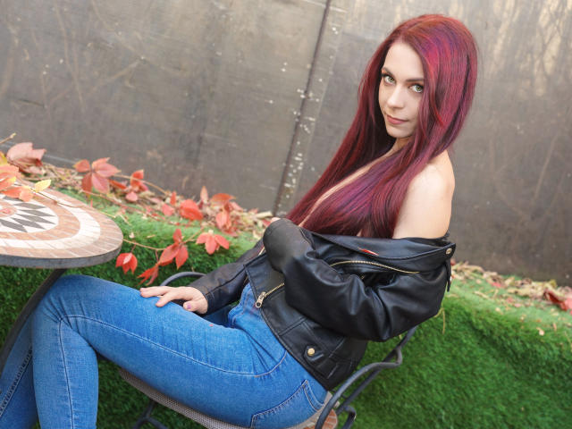 CindeFunRella with red hair, blue jeans, and black leather jacket, off-shoulder, sitting on a green grass bench, looking at camera, small tits, cute, sexy.