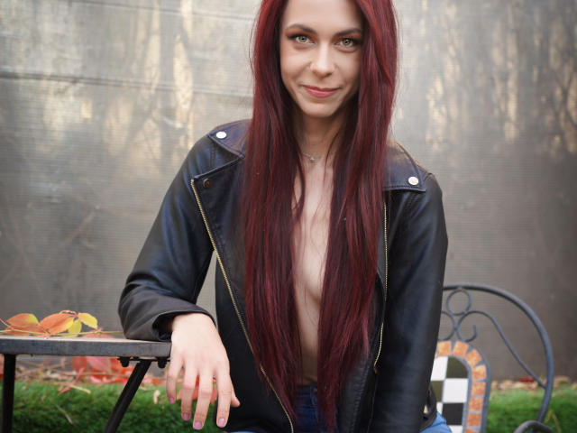 CindeFunRella with long red hair, smiling, wearing a black leather jacket, leaning on a table with autumn leaves, exposing her bare chest. Her hand rests on the table.