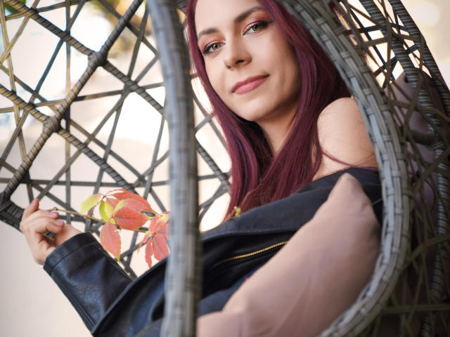CindeFunRella with red hair, pink eyeshadow, and a smile, lounging in a woven hanging chair, wearing a black off-shoulder top, holding a leaf.