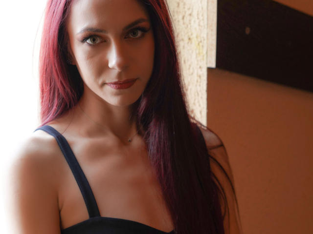 CindeFunRella mit langen, roten Haaren, die ein schwarzes Trägerkleid trägt, hat einen ernsten Gesichtsausdruck, eine mittelgroße Brust, blasse Haut und ein kleines Nasenpiercing und steht vor einer Wand..