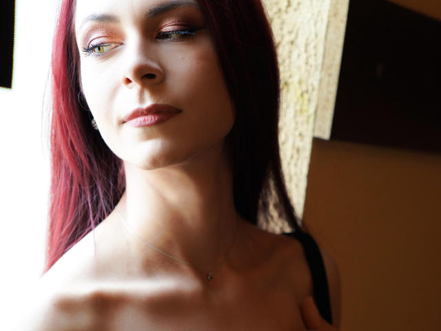 CindeFunRella with red hair, pale skin, and subtle makeup, wearing a black strap, has a small necklace and a slight smile, looking to the side in a soft light.