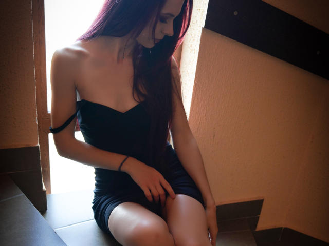 CindeFunRella, con el pelo largo y morado, sentada, vestida con un vestido negro sin tirantes, mostrando sus pequeños pechos y su trasero desnudo, con las manos sobre el muslo, en una luz tenue..