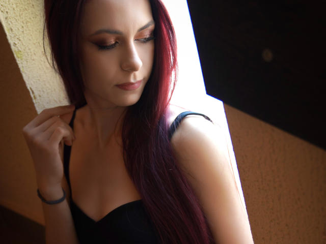 CindeFunRella with red hair, black strap top, and subtle makeup, holding strap, eyes down, soft light, slight smile, wrist bracelet, light skin, textured wall.
