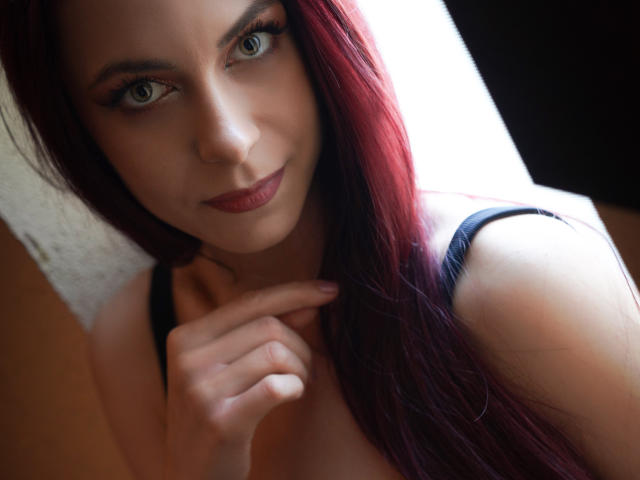 CindeFunRella, con i capelli rossi, gli occhi azzurri e il rossetto scuro, indossa una canotta nera e tocca delicatamente il mento, sorridendo leggermente. Il suo seno è parzialmente visibile..
