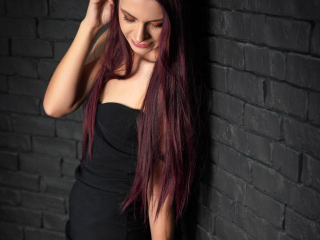 CindeFunRella has long, dark purple hair, wearing a strapless black dress, standing against a black brick wall, smiling with eyes closed, right hand in her hair.