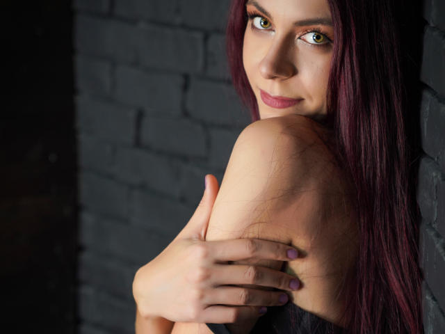 CindeFunRella with red hair, green eyes, and pink lipstick, hugging her bare shoulder, showing off her nipple, against a dark brick wall, smiling subtly.