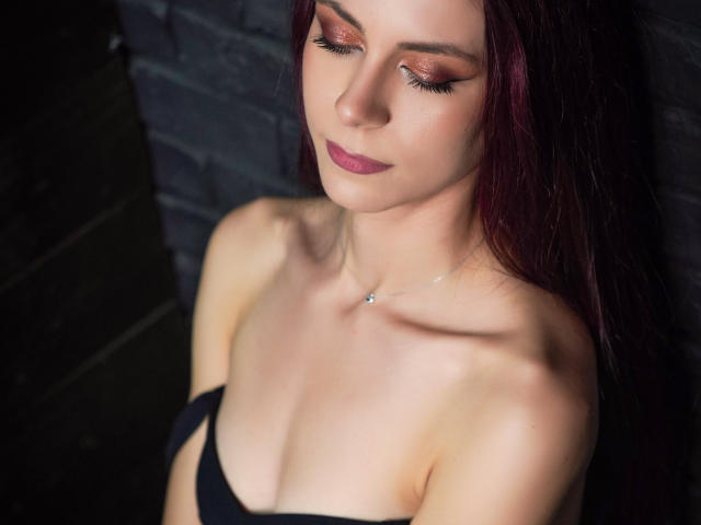 CindeFunRella with red hair, fair skin, and purple lipstick, eyes closed, wearing black strapless top, showing her small tits and collarbone, against dark brick wall.