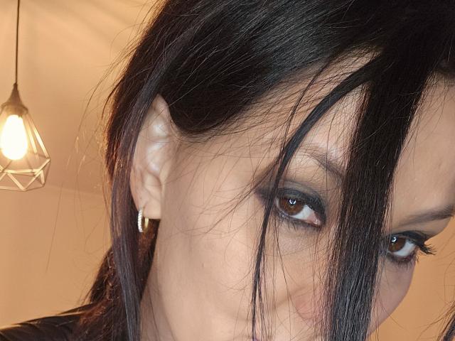 O rosto de JoliDanielle, mostrando seus olhos castanho-escuros com maquiagem esfumada, cabelo preto e um pequeno brinco de prata, com uma luz suave e quente ao fundo..