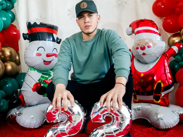 SebastiianMaxime, portant une chemise verte et une casquette noire, est assis entre deux personnages gonflables de Noël, avec les mains sur des ballons rouges et argentés brillants en forme de "20"..