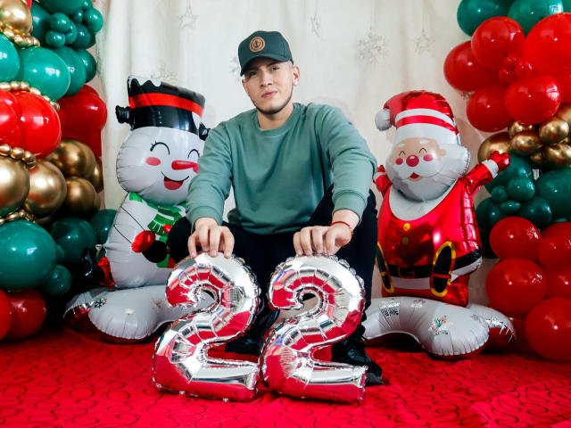 SebastiianMaxime ajoelhado, vestindo uma camisa verde, boné preto e segurando um conjunto de balões vermelhos. Ele está segurando balões brilhantes com o número "22" entre um boneco de neve inflável e um Papai Noel inflável. Tapete vermelho, balões festivos..