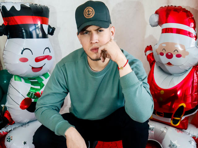 SebastiianMaxime sits in front of inflatable Christmas figures, wearing a green cap, green sweater, and black pants, with a thoughtful expression and hand on chin.