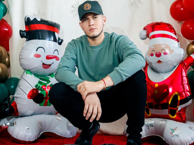SebastiianMaxime est accroupi entre deux Pères Noël gonflables, portant un pull vert, un pantalon noir et une casquette verte, avec un bracelet rouge à son poignet droit..