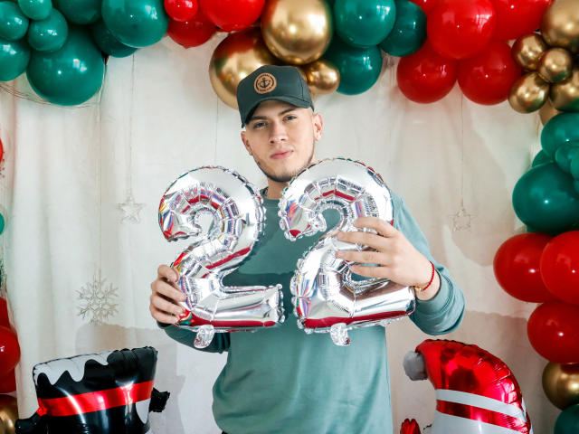 SebastiianMaxime segurando balões prateados brilhantes com o número "21", vestindo uma camisa verde e um boné preto, cercado por balões vermelhos, verdes e dourados, com um chapéu de Papai Noel no canto inferior esquerdo..
