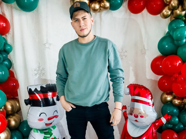 SebastiianMaxime está em frente a balões de Natal, vestindo um suéter verde, calças pretas e um boné preto, com dois bonecos de neve infláveis de cada lado. Sua mão está no bolso..