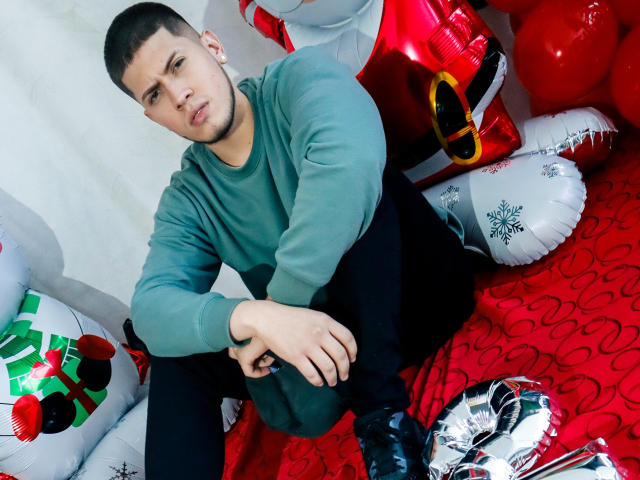SebastiianMaxime, con la cabeza rapada, un suéter verde, pantalones negros y botas negras, está sentado entre globos navideños y regalos, luciendo ligeramente aburrido. Su trasero es parcialmente visible..
