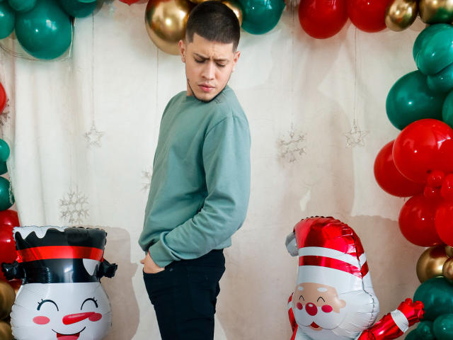 SebastiianMaxime, avec ses cheveux bruns courts, son pull vert et son pantalon noir, une main dans la poche, l'air contrarié, se tient devant des ballons rouges et verts, ainsi que des figurines gonflables sur le thème de Noël..