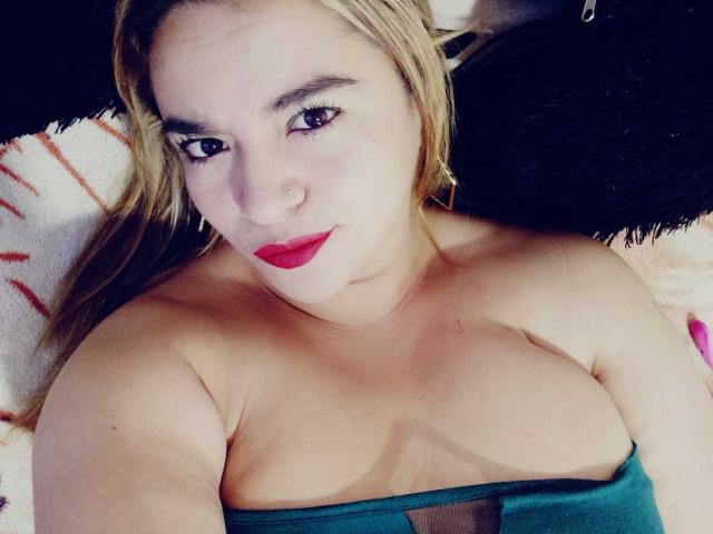 NaugthyCurvy met blond haar, rode lippenstift en een groene top zonder bandjes, waarbij haar borsten zichtbaar zijn, liggend op een zwart kussen, kijkend naar de camera met een lichte glimlach..