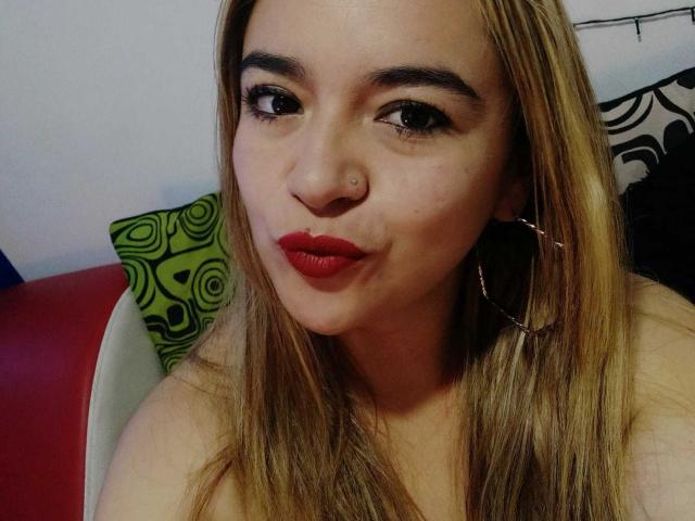 NaugthyCurvy, avec du rouge à lèvres, un piercing au nez et des boucles d'oreilles créoles, a de longs cheveux blonds, une peau claire et regarde l'appareil photo avec un léger sourire. Ses seins sont partiellement visibles..