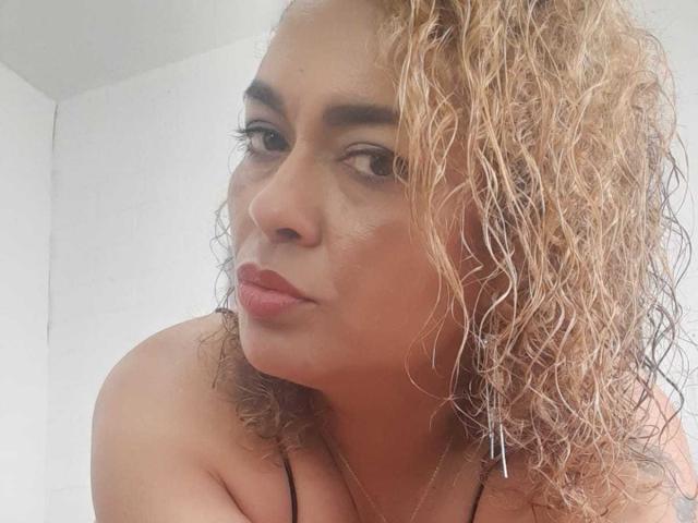SolmaryHot has curly blonde hair, tan skin, and is looking sideways with pouty red lips. She's topless with a black strap, and her tits are partially visible.