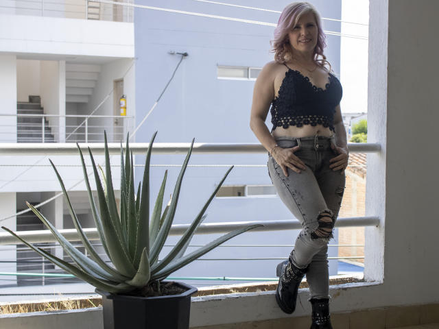 Tina, con i capelli rosa, una maglia di pizzo, jeans strappati e stivali neri, appoggiata al balcone, accanto a una grande pianta succulenta verde. Il seno è visibile, i glutei sono leggermente nascosti dai jeans..