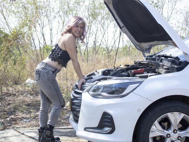 TinaCharter with pink hair, black crop top, gray jeans, black boots, bending over, white car, hood up, smiling, ass in jeans, standing by car in outdoor setting.