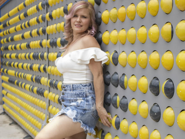 TinaCharter ha i capelli rosa e ricci, indossa una maglietta bianca a spalle scoperte e pantaloncini di jeans sfilacciati, ed è appoggiata a un muro giallo e nero con protuberanze circolari. Il suo sedere è leggermente visibile..