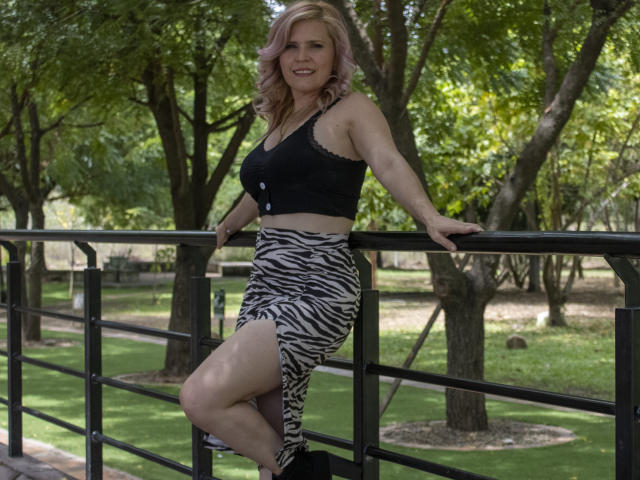 TinaCharter, con i capelli biondi, una maglia di pizzo nera e una gonna a righe zebrate, si appoggia a una ringhiera nera, mostrando il sedere e sorridendo. Si trova in un parco e indossa scarpe nere..