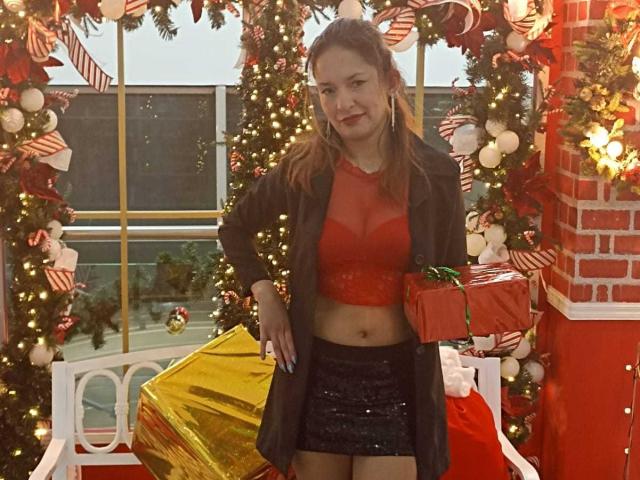 Angela Branson mit rotem Crop-Top, schwarzem paillettenbesetztem Rock, schwarzem Mantel, hält ein rotes Geschenk, steht in einer weihnachtlich gestalteten Umgebung, lächelt, hat hellbraunes Haar, mittelgroße Brüste, ist hübsch..