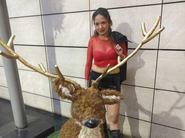Angela Branson met grote borsten, een rode top van gaas, een zwarte rok, zwarte laarzen, staand naast een pluizig hert met gouden hoorns, op een foto..