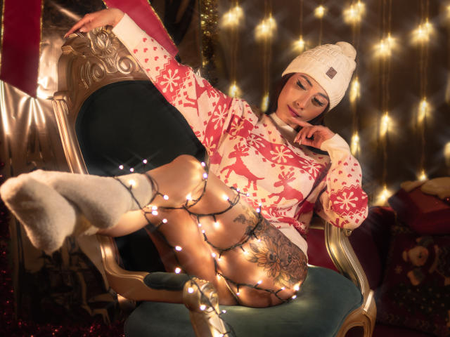 AnyaJay with white beanie, pink reindeer sweater, white socks, and string lights wrapped around her legs and arms, lounging on an ornate chair, showing her tattooed pussy.