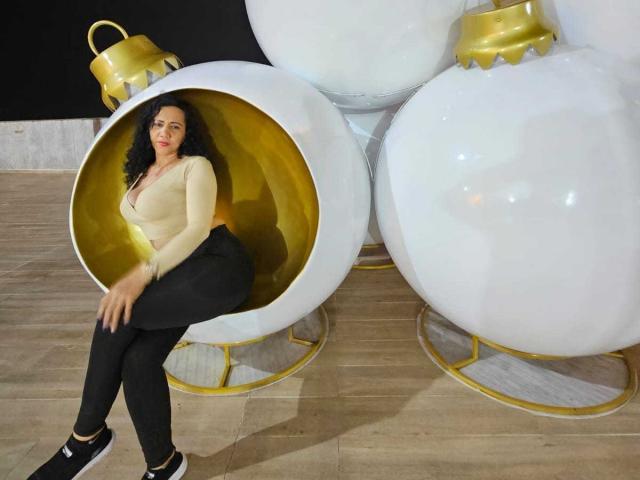 NaomyHott69, avec ses cheveux noirs bouclés, son haut beige, son pantalon noir et ses chaussures noires, est assise dans un fauteuil doré en forme d'œuf, à côté de deux ornements blancs avec des détails dorés..