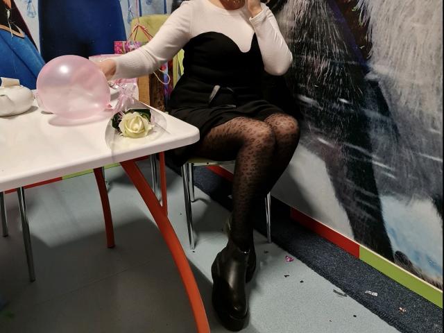 MonaCool portant une robe noire, des manches blanches, des collants à motifs léopard et des talons noirs épais, tenant un ballon rose, assise à une table avec un plateau blanc, une rose et un sac rose..