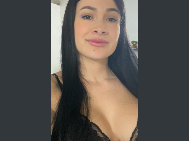 CherryLoux with long black hair, pink lipstick, and purple eyeliner, wearing a black lace top, showing her tits, and a small mole on her chin, smiling at the camera.