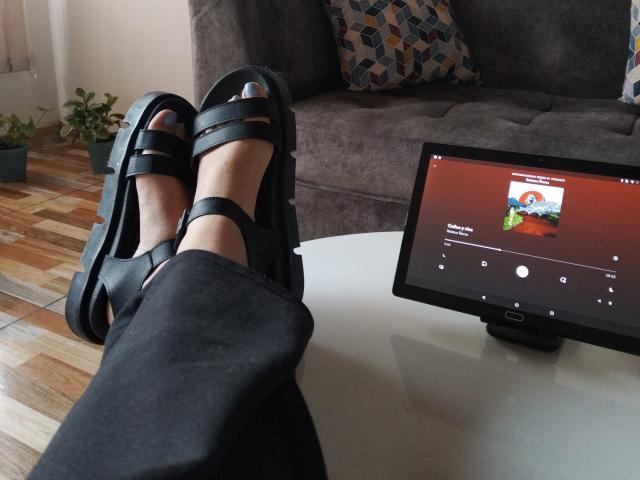 Mllevv's feet in black high-heeled sandals with straps, legs in black pants, resting on a white table, tablet showing a video with a woman's face in front.