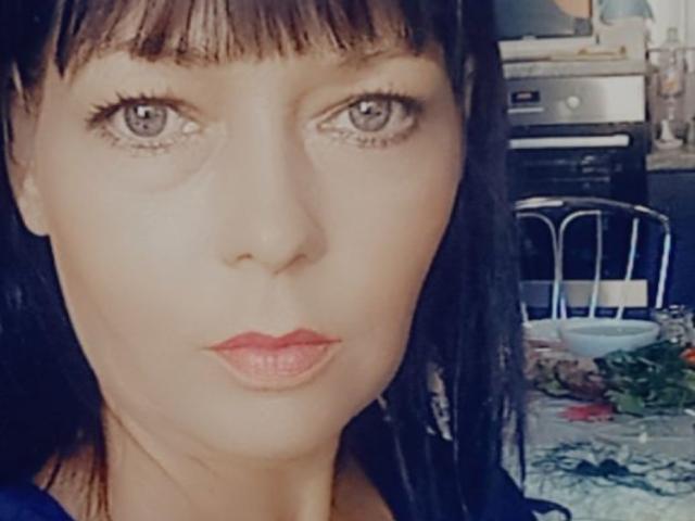 IlonaKristie heeft bleke huid, donker steil haar met een pony, grijze ogen en rode lippenstift. Ze kijkt recht in de camera met een serieuze uitdrukking..