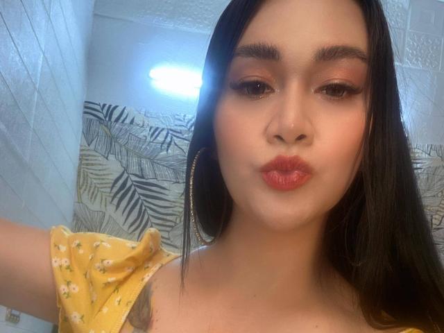 AvirySins with pouty red lips, brown eyes, and long black hair, wearing a yellow floral top, large hoop earrings, and a subtle tattoo on her left shoulder.