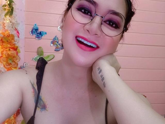 AvirySins met roze make-up, een bril en roze lippenstift, lachend met tanden zichtbaar, hand op de wang, een tatoeage op de pols, een zwarte band, kleurrijke vlindertatoeages en een roze muur..