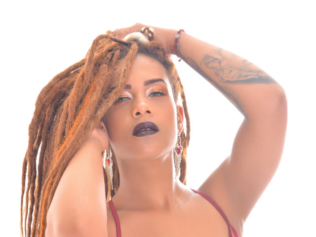 KendallQuinn with orange dreadlocks, dark lipstick, and tattoos, hand in hair, wearing red earrings and bra, white background, looking up with a confident expression.