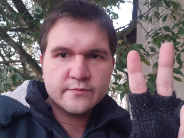 NectarineFad, a chubby guy with short brown hair, wearing a black jacket and fingerless gloves, showing a peace sign with his right hand, has a slight scar on his forehead.