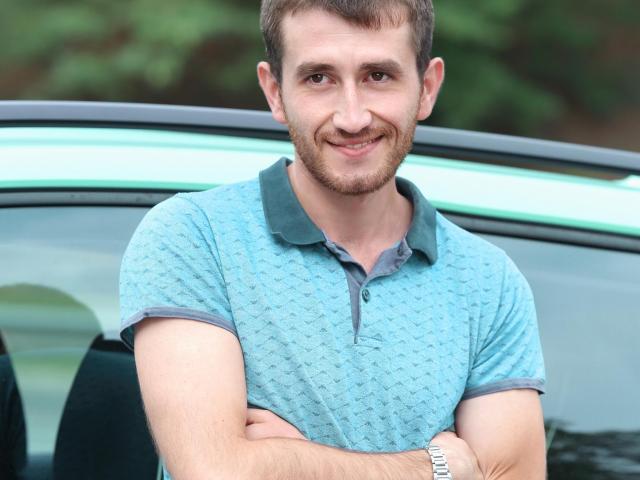 TastyAndrew, a cute dude with short brown hair and a beard, wearing a blue patterned polo, arms crossed, smiling at the camera, standing by a green car.