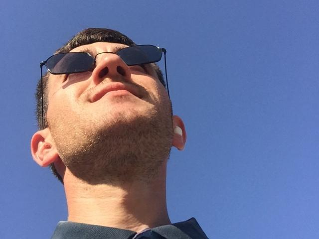 TastyAndrew, com um leve sorriso, usando óculos de sol pretos, olhando para o céu azul claro. Ele tem uma barba curta e parece estiloso..