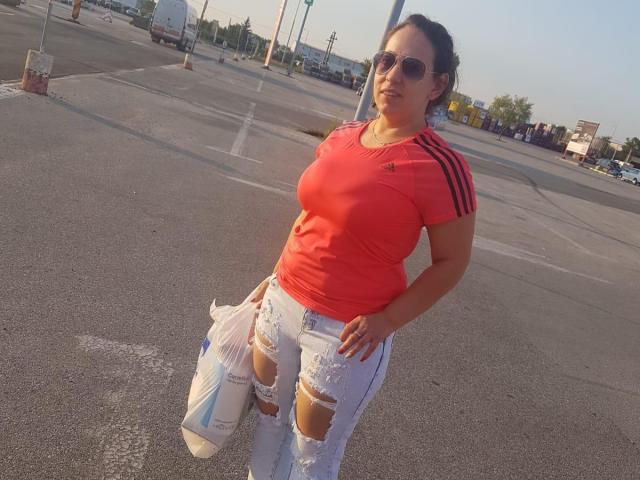 RenattaRosse está de pie en un estacionamiento, vestida con una camiseta deportiva roja ajustada, jeans blancos rotos y zapatillas blancas, con una mano en la cadera y gafas de sol redondas..