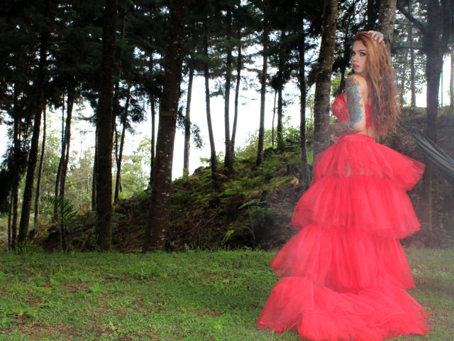 KattyCandyX with long red hair, wearing a red, layered, tulle dress, tattooed arms, standing in a forest, looking back, green grass, and tall trees in the background.