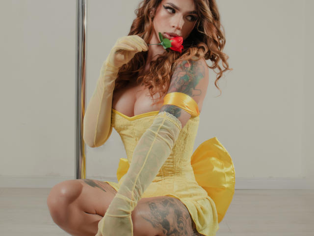 KattyCandyX, con cabello castaño ondulado, vestido amarillo y guantes largos, sosteniendo una rosa roja, mostrando tatuajes en su brazo y muslo, y sus grandes senos..