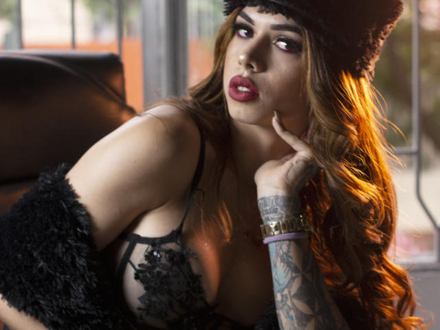 KattyCandyX with long brown hair, red lipstick, black lace bra, tattoos on right arm, black furry hat, and black fur coat, looking seductively at camera.