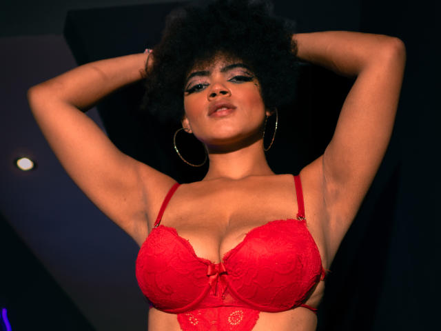 Alice Jelanni, con los brazos levantados, vistiendo un sujetador de encaje rojo, mostrando sus grandes senos, piel oscura, peinado afro, aros de oro y maquillaje llamativo en una habitación con poca luz..